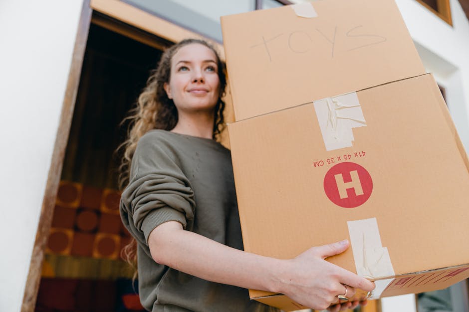 A young woman with dark hair, wearing a white T-shirt, sits on the floor of a residential property during a home relocation process, handling packing materials and documents. The room behind her is fi